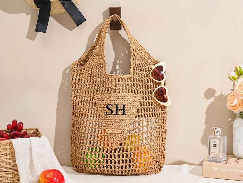 May include: A woven straw tote bag with a large open weave design. The bag has a pocket with the initials "SH" and is hanging on a wooden hook. Heart-shaped sunglasses are attached to the bag. A straw hat with a black bow hangs above.