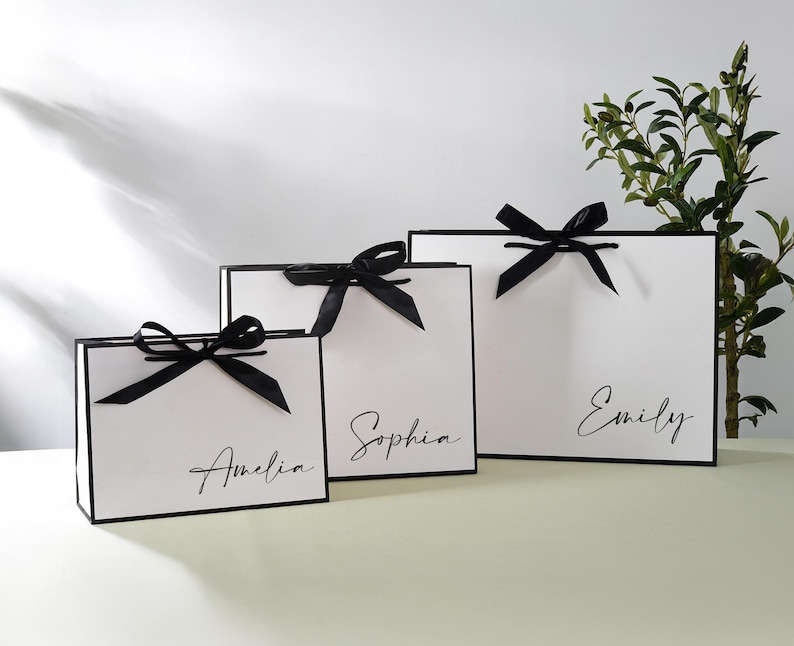 May include: Three white gift bags with black ribbon bows and personalised names. The bags are arranged in a row from largest to smallest, with the names "Emily", "Sophia", and "Amelia" written in black script.