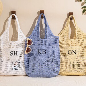 May include: Three open-weave tote bags in white, blue, and beige hang on wooden pegs. Each bag has a pocket with initials: "SH", "KB", and "GN". Sunglasses rest on the blue bag. A straw hat and flowers are also visible.