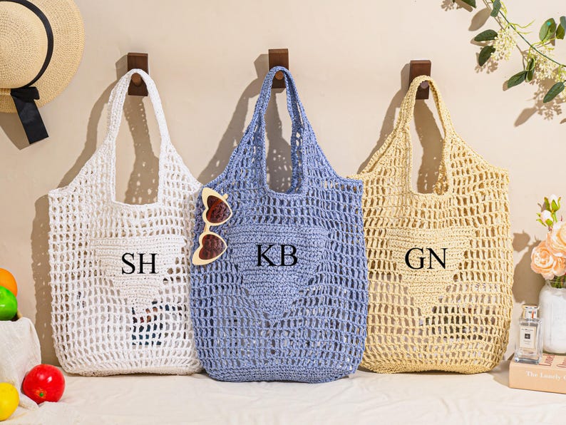 May include: Three open-weave tote bags in white, blue, and beige hang on wooden pegs. Each bag has a pocket with initials: "SH", "KB", and "GN". Sunglasses rest on the blue bag. A straw hat and flowers are also visible.