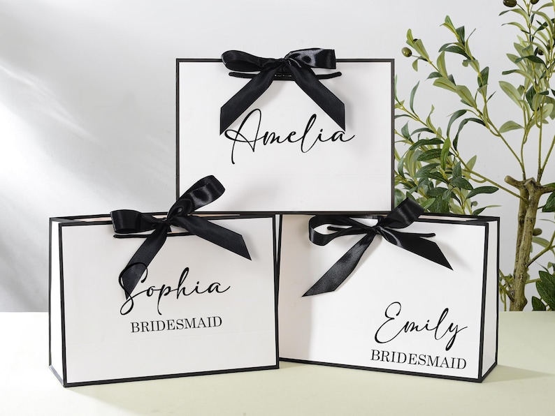 May include: Three white gift bags with black ribbon bows and black trim. The bags are personalised with the names "Amelia", "Sophia Bridesmaid", and "Emily Bridesmaid".
