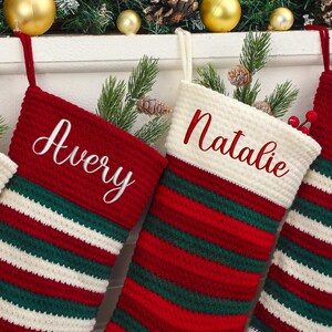 May include: Close-up of personalized Christmas stockings. The stockings are red, white, and green striped with the names "Avery" and "Natalie" embroidered in white script. The stockings are hanging with festive decorations.