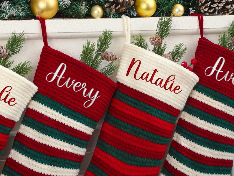 May include: Close-up of personalized Christmas stockings. The stockings are red, white, and green striped with the names "Avery" and "Natalie" embroidered in white script. The stockings are hanging with festive decorations.