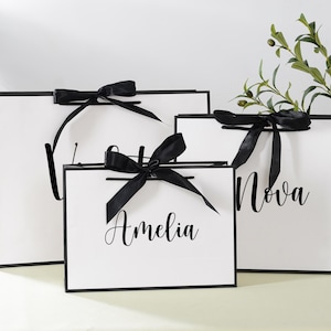 May include: Three white gift bags with black handles and black ribbon bows. The bags are labelled with the names "Amelia", "Nova", and "Bridesmaid".
