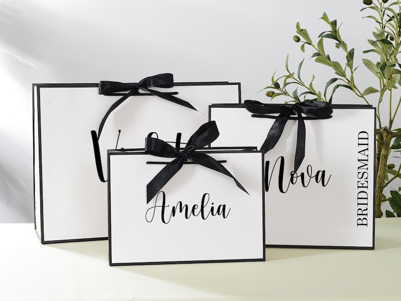 May include: Three white gift bags with black handles and black ribbon bows. The bags are labelled with the names "Amelia", "Nova", and "Bridesmaid".