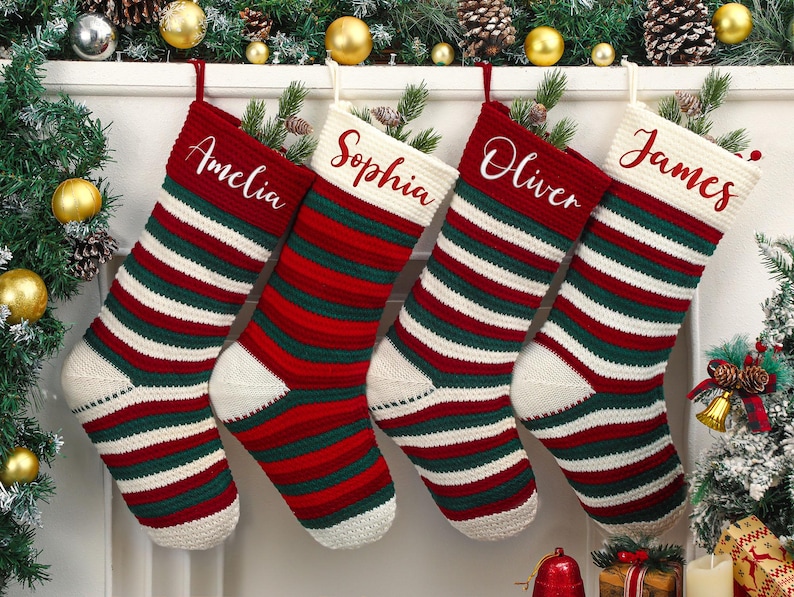 May include: Four Christmas stockings with red, green, and white stripes hang from a white mantel. Each stocking has a name embroidered in cursive: Amelia, Sophia, Oliver, and James. The stockings are decorated with pine and gold ornaments.
