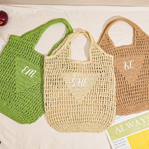 May include: Three crocheted tote bags in green, beige, and tan. Each bag has a heart-shaped pocket with white embroidered initials. The bags are open-weave and have a handle for carrying. A newspaper with the words "ALWAYS FLOW" is visible.
