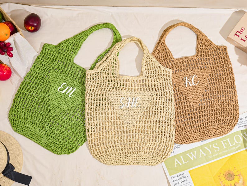 May include: Three crocheted tote bags in green, beige, and tan. Each bag has a heart-shaped pocket with white embroidered initials. The bags are open-weave and have a handle for carrying. A newspaper with the words "ALWAYS FLOW" is visible.