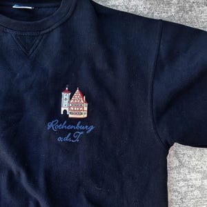 Può includere: Una felpa girocollo blu navy con un ricamo raffigurante un edificio a graticcio e una torre dell'orologio. Sotto il ricamo, il testo Rothenburg o.d.T. è cucito con filo blu. Il capo presenta un colletto a coste con un inserto a V.
