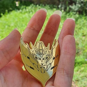 May include: A gold-coloured, shield-shaped pin featuring a detailed, stylised face design. The pin has a symmetrical design with sharp angles and black accents, held in a hand against a blurred green background.