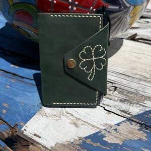 May include: A dark green leather notebook cover featuring a hand-stitched four-leaf clover design on the front flap. The cover is secured with a single brass-colored metal snap button and includes a black pen tucked into the side loop.