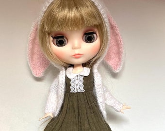 Op de afbeelding: Een pop met blond haar die een wit gebreid vestje draagt met roze konijnenoren, een groene jurk en witte schoenen.