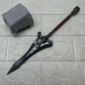 Puede incluir: Una espada de fantasía gris oscuro detallada con una empuñadura decorativa y un mango envuelto en marrón. La hoja es larga y puntiaguda. Un objeto gris cubierto de tela está en el fondo.