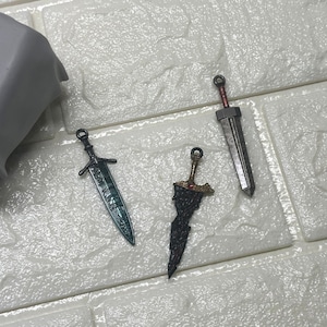 Puede incluir: Tres espadas en miniatura con diseños detallados. Una espada tiene una hoja verde, otra una hoja negra con detalles dorados y la tercera una hoja plateada con un mango rojo. Las espadas se exhiben sobre una superficie blanca texturizada.