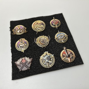 May include: A collection of nine ornate, gold-colored metal pins with various designs, including a dragon, a skull, and crossed axes. Each pin has intricate details and is set against a black background.