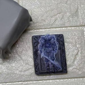 Peut inclure: Une tuile décorative rectangulaire gris foncé avec une illustration détaillée d'un chevalier en armure, rendu dans des tons de bleu. Le chevalier tient une arme lumineuse. La tuile a une bordure ornée.