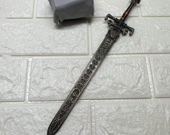 May include: A detailed, silver-colored miniature sword with an ornate handle and blade. The handle features a dark red grip and intricate cross-shaped design. The blade is engraved with floral patterns and crosses. A gray, cloth-covered object is in the background.