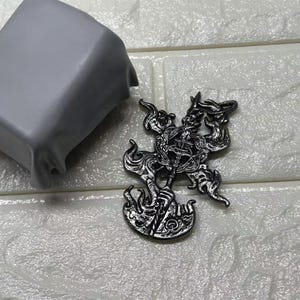 May include: A detailed, silver-colored metal pendant with an intricate design featuring a central figure surrounded by swirling patterns and flames. The pendant has a dark, antique finish, and is likely a piece of jewelry.