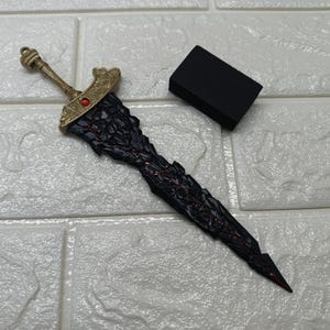 Könnte beinhalten: Ein detailliertes Fantasy-Schwert mit schwarzer Klinge und goldenem Knauf. Die Klinge hat ein strukturiertes, gezacktes Design mit roten Akzenten. Der Knauf hat einen roten Edelstein. Ein kleines schwarzes rechteckiges Objekt befindet sich neben dem Schwert.