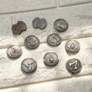 Könnte beinhalten: Eine Sammlung von zehn runden Metallknöpfen mit verschiedenen eingravierten Symbolen, darunter das Assassin's Creed-Logo, ein Auge des Horus, ein Totenkopf und ein roter Stern. Die Knöpfe haben eine silberne Metalloberfläche mit texturiertem Hintergrund.