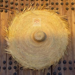 May include: A large, round straw hat with a wide brim and a central crown. The hat is made of woven straw and has a natural, light brown color. The brim has a fringed edge. The hat is resting on a wooden surface.