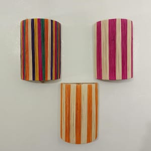 Könnte beinhalten: Drei dekorative, zylindrische Wandpflanzgefäße. Eines hat vertikale Streifen in Orange, Gelb, Grün, Pink und Lila. Ein weiteres hat rosa und weiße Streifen, und das dritte hat orange und weiße Streifen. Alle sind an einer weißen Wand befestigt.