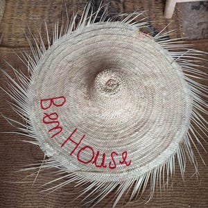 May include: A round, woven straw hat with a conical crown and a wide brim. The hat features the embroidered text "Ben House" in red. The brim is fringed with long, thin straw pieces.
