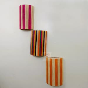 May include: Three semi-cylindrical wall sconces featuring vertical striped patterns. The top sconce has pink and natural tan stripes, the middle has purple, green, orange, and tan stripes, and the bottom has orange and natural tan stripes.