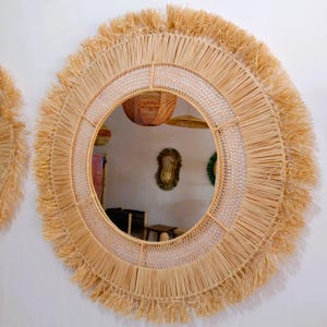 May include: A round wall mirror with a natural woven raffia frame. The mirror has a fringe detail around the outer edge and a woven pattern in the middle. The mirror is hanging on a white wall.