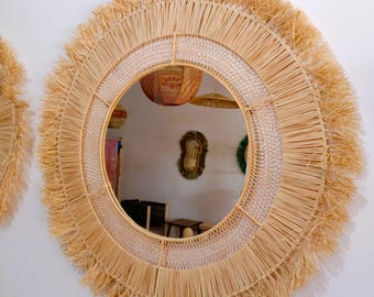 May include: A round wall mirror with a natural woven raffia frame. The mirror has a fringe detail around the outer edge and a woven pattern in the middle. The mirror is hanging on a white wall.