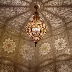Pode incluir: Luminária pendente dourada e ornamentada com intrincados recortes florais, projetando sombras decorativas no teto e nas paredes. A lâmpada está iluminada, criando um ambiente acolhedor e convidativo. O design lembra a decoração marroquina ou do Oriente Médio.