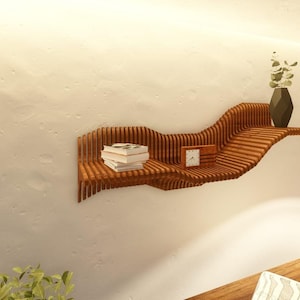 Op de afbeelding: Een modern houten wandrek met een uniek, golvend ontwerp. Het rek bevat een stapel boeken, een kleine klok en een donkere vaas met groen. Het rek is bevestigd aan een getextureerde witte muur.
