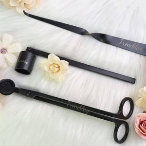 Puede incluir: Un conjunto de accesorios para velas negros, que incluyen un sumergidor de mecha, un recortador de mecha y un apagador, dispuestos sobre una superficie blanca y esponjosa. La palabra "Tremblay" está inscrita en cada herramienta. Flores decorativas rodean las herramientas.