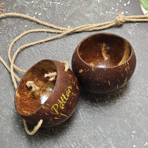 以下が含まれることがあります： 自然な茶色の仕上げが施された、2つの手作りのココナッツシェルボウル。1つのボウルには、金色の文字で「Patton」という名前が書かれています。両方のボウルは天然のロープでつながっています。ボウルは装飾用または機能的な使用を目的としている可能性があります。