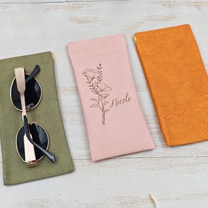 May include: Three eyeglass cases in olive green, pink, and orange. The olive green case holds round sunglasses with gold frames. The pink case has a floral design and the name "Nicole". The orange case is plain.