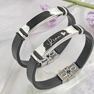 May include: Two black leather bracelets with silver-tone metal accents. One bracelet has the name "Lisa" engraved with a heart symbol. The bracelets have a clasp closure and are displayed on a white surface.