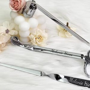 Pode incluir: Um conjunto de três acessórios de vela prateados: um mergulhador de pavio, um aparador de pavio e um abafador. O aparador e o abafador têm o nome "Melanie Hiess" gravado. O mergulhador de pavio tem o nome "Dana" gravado.