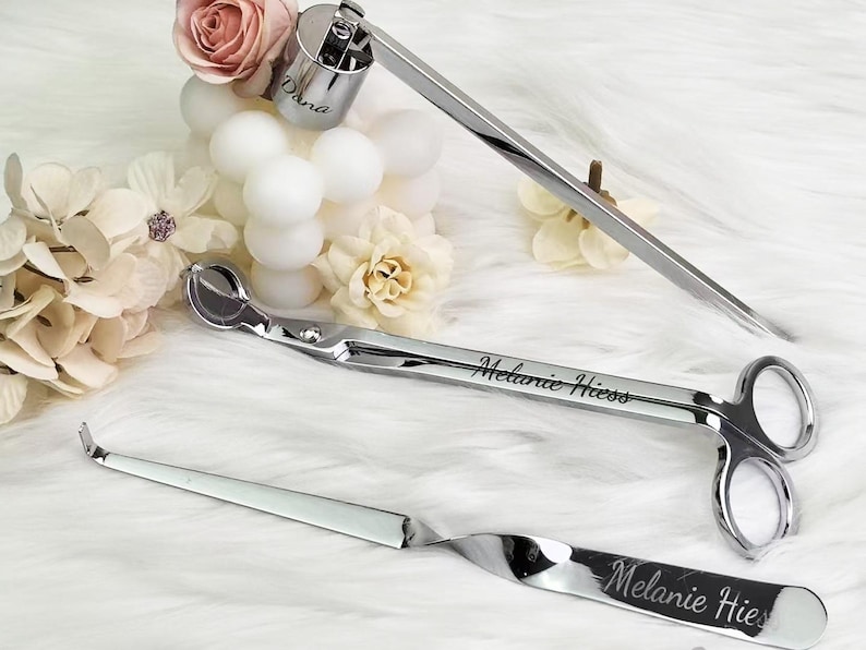 Op de afbeelding: Een set van drie zilveren kaarsenaccessoires: een lontdipper, een lonttrimmer en een snuffer. De lonttrimmer en snuffer zijn gegraveerd met de naam "Melanie Hiess". De lontdipper heeft de naam "Dana" gegraveerd.