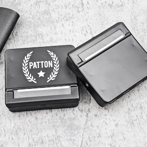 May include: Two black rolling machines are displayed on a gray surface. One is open, revealing a silver roller. The other is closed, with the name "PATTON" in white, surrounded by a laurel wreath and a star.