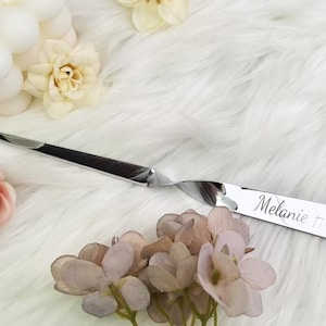 Op de afbeelding: Een glanzende, zilveren briefopener met een gebogen lemmet en een gedraaide handgreep. De naam "Melanie" is op de handgreep gegraveerd. De briefopener ligt op een witte, pluizige ondergrond met bloemaccenten.