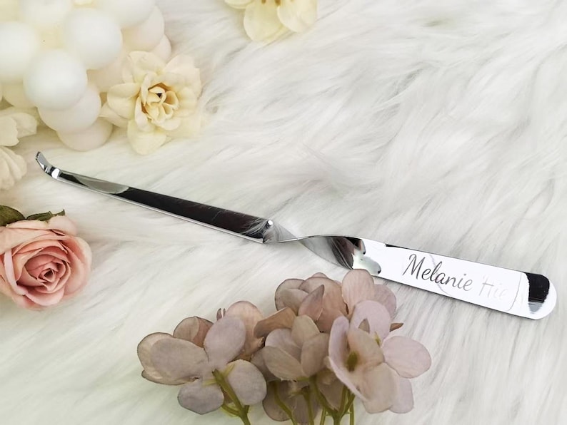 Op de afbeelding: Een glanzende, zilveren briefopener met een gebogen lemmet en een gedraaide handgreep. De naam "Melanie" is op de handgreep gegraveerd. De briefopener ligt op een witte, pluizige ondergrond met bloemaccenten.