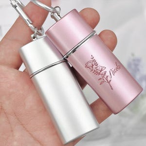 Op de afbeelding: Twee cilindrische metalen containers met sleutelhangers. Eén is zilverkleurig, de andere roze en voorzien van een bloemenontwerp en de gegraveerde naam "Nicole". Beide hebben een zilveren band in het midden.