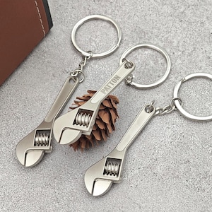 Peut inclure: Trois porte-clés en forme de clé à molette argentés avec des anneaux. L'une des clés porte l'inscription "PATTON". Les porte-clés sont présentés sur une surface grise avec un objet marron en arrière-plan.