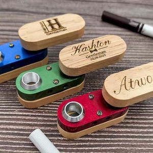 Puede incluir: Una colección de pitilleras personalizadas de madera en azul, verde y rojo, cada una con una tapa de madera a juego. Las tapas están grabadas con nombres y la palabra "Gentleman".