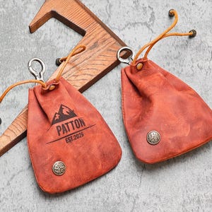 May include: Two rust-colored leather pouches with drawstring closures and metal accents. One pouch has the name "PATTON" and "EST.2026" printed on it, along with a mountain graphic. The pouches are designed to hold small items.