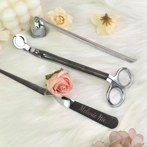 Op de afbeelding: Een set kaarsenaccessoires, waaronder een lonttrimmer, een snuffer en een dipper, gerangschikt op een witte, donzige ondergrond. De tools zijn zilver en zwart, met de naam "Melanie Hiess" gegraveerd op twee ervan. Decoratieve bloemen omringen de tools.