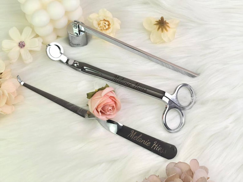 Op de afbeelding: Een set kaarsenaccessoires, waaronder een lonttrimmer, een snuffer en een dipper, gerangschikt op een witte, donzige ondergrond. De tools zijn zilver en zwart, met de naam "Melanie Hiess" gegraveerd op twee ervan. Decoratieve bloemen omringen de tools.