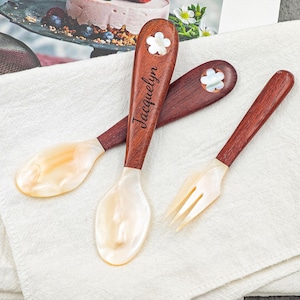 Puede incluir: Tres utensilios de servir de madera con cabezas de nácar. Una cuchara está grabada con el nombre "Jacquelyn". Los mangos son de color marrón oscuro con una incrustación en forma de flor. Los utensilios están sobre un paño blanco.