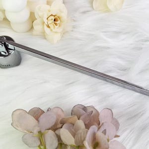 Op de afbeelding: Een zilveren kaarsenblusser met een lange, rechthoekige handgreep en een ronde basis. De basis heeft het woord "ana" gegraveerd. De blusser ligt op een wit, pluizig oppervlak, met bloemen op de achtergrond.