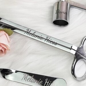 Op de afbeelding: Een set zilveren kaarsaccessoires, waaronder een lonttrimmer, een snuffer en een kaarshouder. De lonttrimmer en snuffer zijn gegraveerd met de naam "Melanie Hiess". Een kleine roze roos staat naast de trimmer.